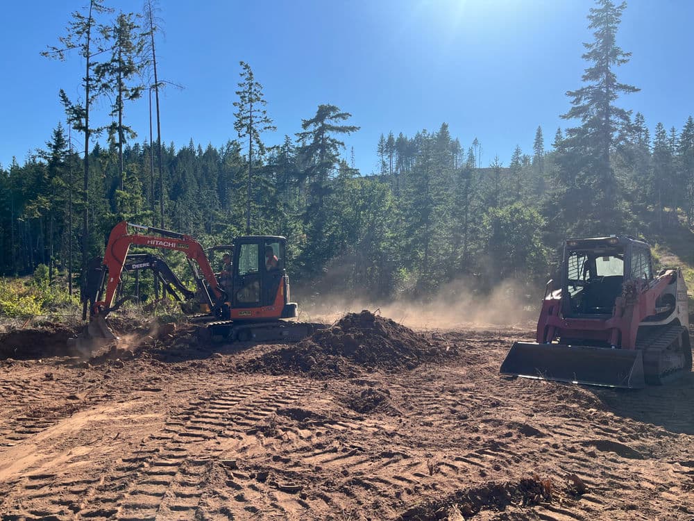 Excavators working on a construction site in a forested area with dust and trees in the background.